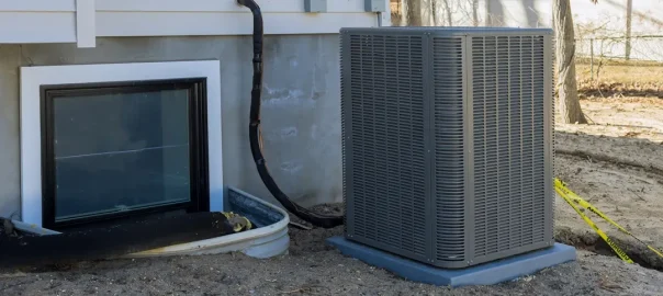 A HVAC system placed over a muddy ground next to the corner of a home and an egress window.