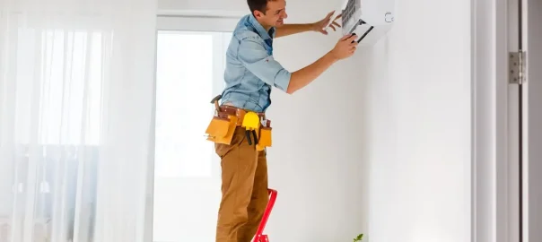 Smiling man stands up on the ladder with his hands placed over the air conditioner unit and mechanic tools hanging all around his waist.